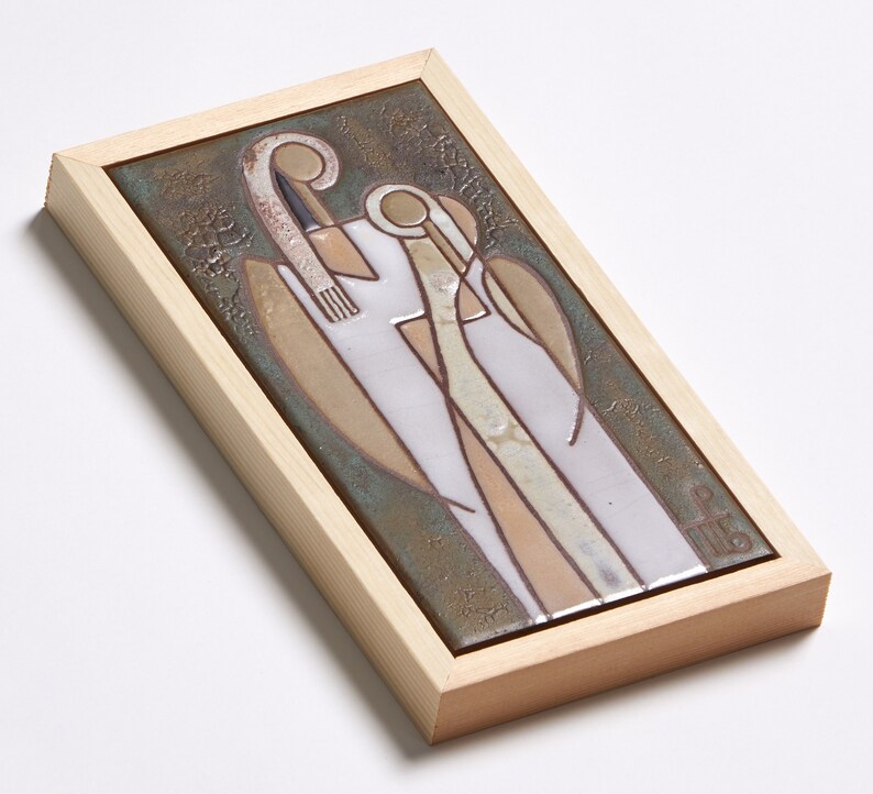 May include: A framed ceramic tile with a stylized image of two figures in white, brown, and beige against a green background. The figures are standing close together and appear to be embracing. The tile is framed in a light wood frame.