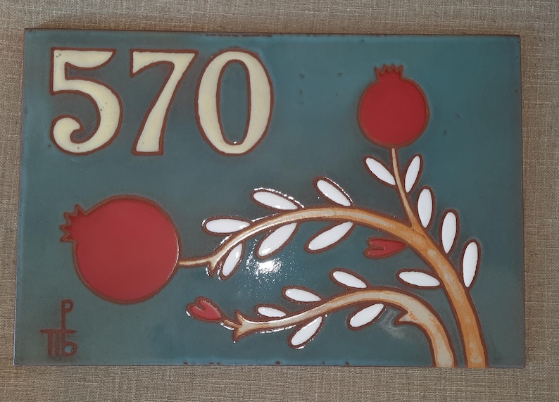 May include: A blue ceramic tile with the number 570 in white and a stylized red pomegranate branch with white leaves.