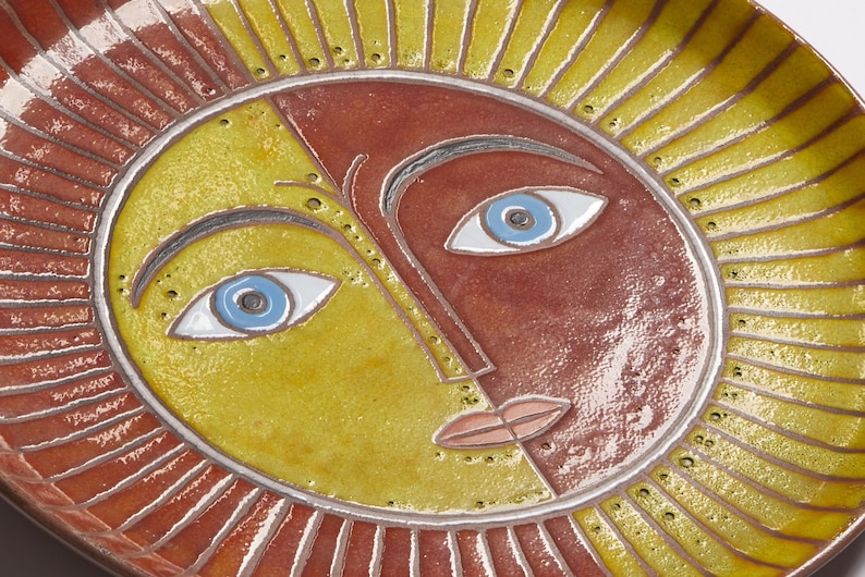 May include: A ceramic plate with a stylized sun face design. The sun has a yellow and red face with blue eyes and a pink mouth. The plate is decorated with a red and yellow sunburst pattern.