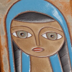 May include: A ceramic tile with a stylized portrait of a person with blue hair and brown skin. The person has large eyes with white pupils and a red mouth.
