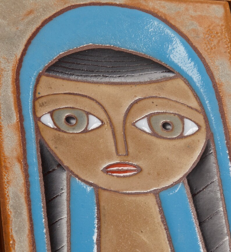 May include: A ceramic tile with a stylized portrait of a person with blue hair and brown skin. The person has large eyes with white pupils and a red mouth.