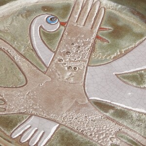 May include: A ceramic plate with a green glaze and a white glaze. The plate features a design of two stylized birds with outstretched wings. The birds are painted in white and have blue eyes.
