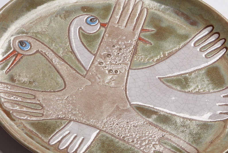 May include: A ceramic plate with a green glaze and a white glaze. The plate features a design of two stylized birds with outstretched wings. The birds are painted in white and have blue eyes.