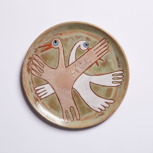 May include: A ceramic plate with a green glaze features a stylized design of two birds with outstretched wings. The birds are painted in shades of white, brown, and orange.