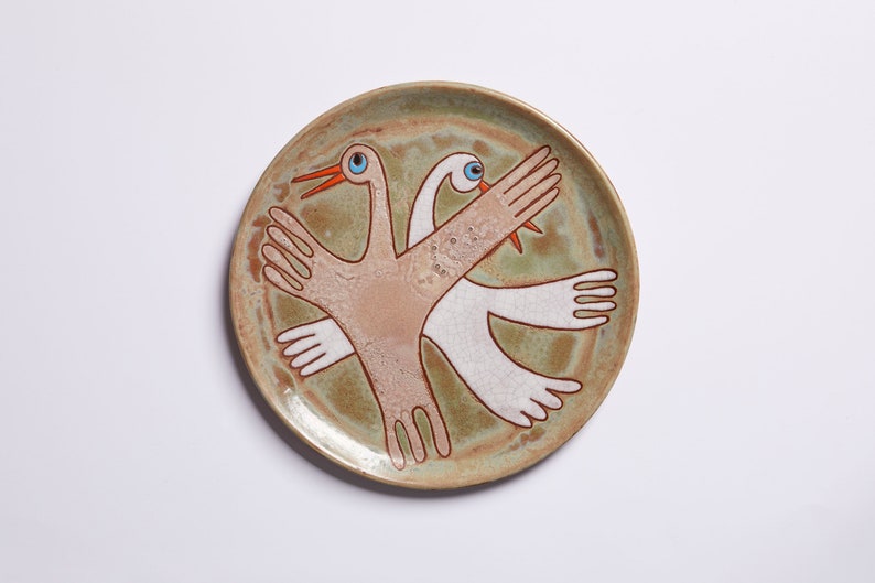 May include: A ceramic plate with a green glaze features a stylized design of two birds with outstretched wings. The birds are painted in shades of white, brown, and orange.