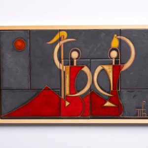 Op de afbeelding: Een ingelijst keramisch tegelkunstwerk met twee gestileerde figuren in rood en goud tegen een donkergrijze achtergrond. De figuren houden voorwerpen vast, een een gele halve maan en de andere een gele peer. Een rode cirkel en een rode berg zijn ook afgebeeld in het kunstwerk.