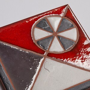 May include: A square ceramic tile with a geometric design. The tile is divided into four sections, each with a different color: red, white, gray, and black. The center of the tile features a white circle with a gray, six-pointed star.