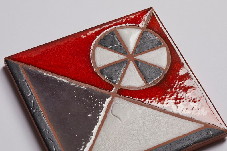 May include: A square ceramic tile with a geometric design. The tile is divided into four sections, each with a different color: red, white, gray, and black. The center of the tile features a white circle with a gray, six-pointed star.