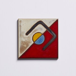 K&ouml;nnte beinhalten: Eine Keramikfliese mit geometrischem Muster. Die Fliese ist in zwei Abschnitte unterteilt, einen roten und einen wei&szlig;en. Der rote Abschnitt hat eine schwarze geometrische Form mit einem gelben und blauen Kreis darin.