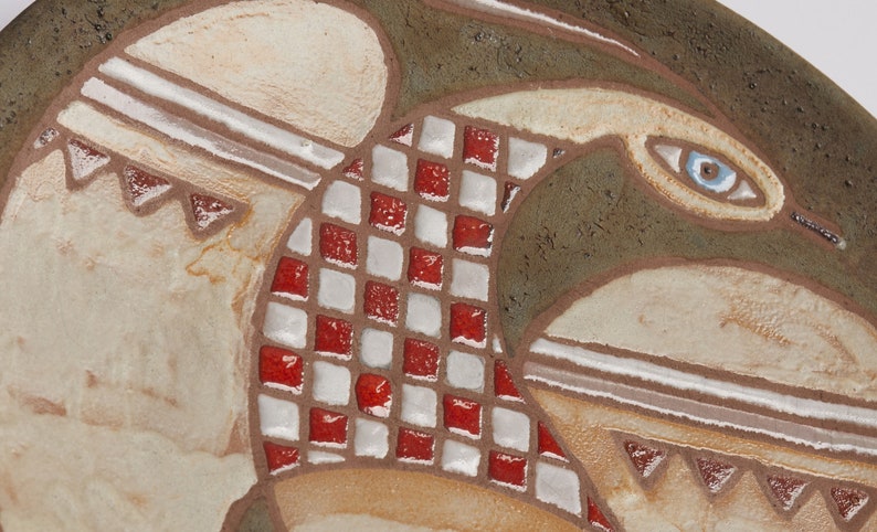 Op de afbeelding: Een keramische wandplaat met een gestileerd vogelontwerp. De vogel is afgebeeld in tinten bruin, wit en rood, met een blauw oog. De plaat heeft een getextureerd oppervlak en een geometrisch patroon.