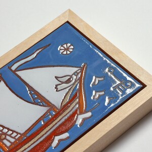 Peut inclure: Un carreau de c&eacute;ramique encadr&eacute; avec un fond bleu repr&eacute;sentant un voilier avec des voiles blanches et une coque rouge. Le carreau est entour&eacute; d'un cadre en bois clair.