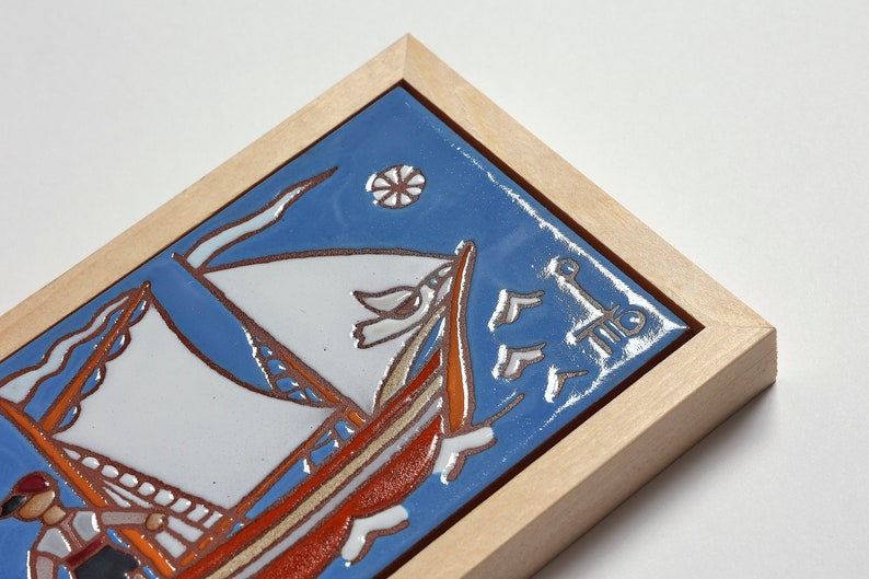 Peut inclure: Un carreau de c&eacute;ramique encadr&eacute; avec un fond bleu repr&eacute;sentant un voilier avec des voiles blanches et une coque rouge. Le carreau est entour&eacute; d'un cadre en bois clair.
