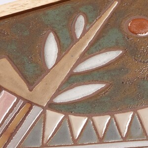 May include: A ceramic tile with a stylized bird design. The bird is pink, brown, and blue with a white eye. The background is a mottled green with a brown circle.