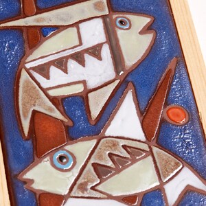 May include: Two stylized fish are depicted on a blue background with brown and white accents. The fish are made of ceramic tiles and are framed in wood.