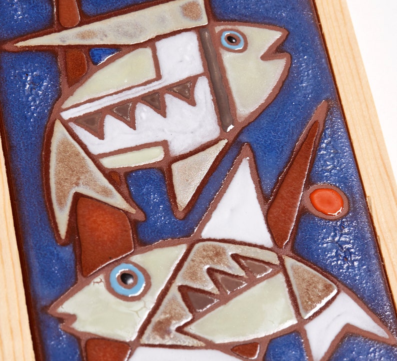 May include: Two stylized fish are depicted on a blue background with brown and white accents. The fish are made of ceramic tiles and are framed in wood.
