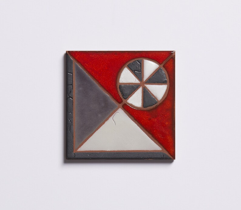 May include: A square ceramic tile with a geometric design. The tile is divided into four sections, each with a different color: red, gray, white, and black. The center of the tile features a black and white circle with a radial design.