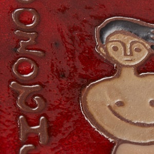 Może przedstawiać: Czerwona płytka ceramiczna z brązowym konturem osoby z uśmiechającą się twarzą. Płytka ma napis "SHOMO" napisany pionowo po lewej stronie.