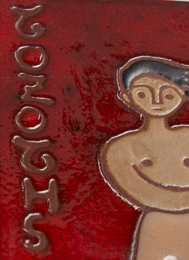 Może przedstawiać: Czerwona płytka ceramiczna z brązowym konturem osoby z uśmiechającą się twarzą. Płytka ma napis "SHOMO" napisany pionowo po lewej stronie.