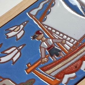 Peut inclure: Un carreau de c&eacute;ramique bleu et blanc avec une sc&egrave;ne peinte d'un voilier avec une voile rouge et blanche, une personne portant un chapeau rouge et des oiseaux blancs volant dans le ciel. Le carreau est encadr&eacute; de bois.