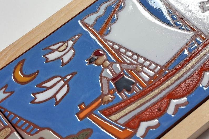 Peut inclure: Un carreau de c&eacute;ramique bleu et blanc avec une sc&egrave;ne peinte d'un voilier avec une voile rouge et blanche, une personne portant un chapeau rouge et des oiseaux blancs volant dans le ciel. Le carreau est encadr&eacute; de bois.