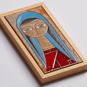 May include: A framed ceramic tile with a colorful illustration of a woman with blue hair and a red dress. The tile is framed in a light wood frame.