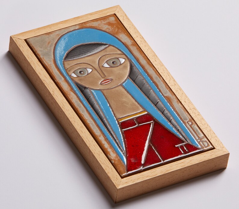 May include: A framed ceramic tile with a colorful illustration of a woman with blue hair and a red dress. The tile is framed in a light wood frame.