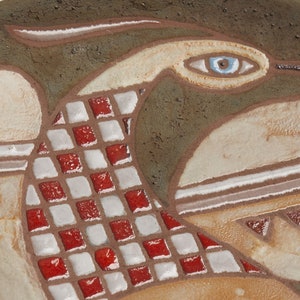 Op de afbeelding: Een keramisch bord met een gestileerd vogelontwerp. De vogel heeft een blauw oog en is omringd door een rood-wit geruit patroon.