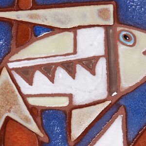May include: A ceramic tile with a stylized image of a fish in shades of brown, white, and blue on a blue background. The fish has a large eye and a wide mouth.