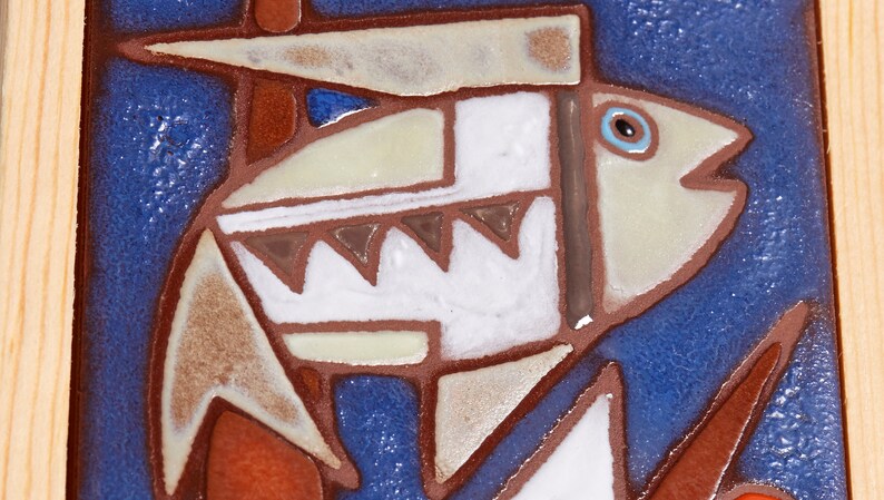 May include: A ceramic tile with a stylized image of a fish in shades of brown, white, and blue on a blue background. The fish has a large eye and a wide mouth.