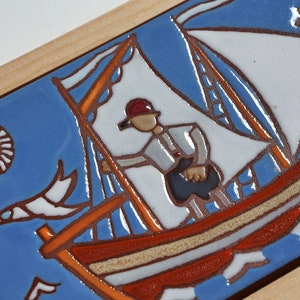 Peut inclure: Un carreau de c&eacute;ramique avec un fond bleu repr&eacute;sentant un voilier avec une voile blanche et une coque rouge et orange. Une personne portant un chapeau rouge se tient sur le bateau. Le soleil est repr&eacute;sent&eacute; comme un cercle rouge avec des rayons blancs. Un oiseau blanc vole dans le ciel.