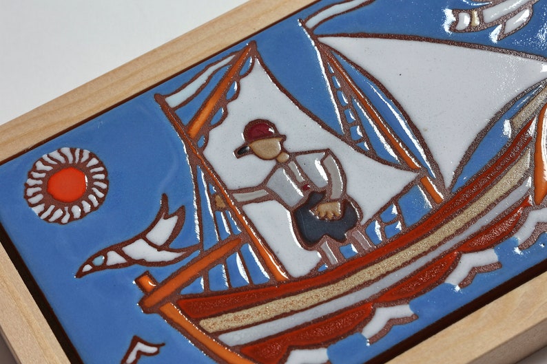Peut inclure: Un carreau de c&eacute;ramique avec un fond bleu repr&eacute;sentant un voilier avec une voile blanche et une coque rouge et orange. Une personne portant un chapeau rouge se tient sur le bateau. Le soleil est repr&eacute;sent&eacute; comme un cercle rouge avec des rayons blancs. Un oiseau blanc vole dans le ciel.