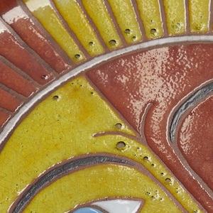 May include: Close-up of a ceramic sun face with yellow, red, and blue paint. The sun has a stylized face with two eyes and a nose. The sun is surrounded by yellow rays.