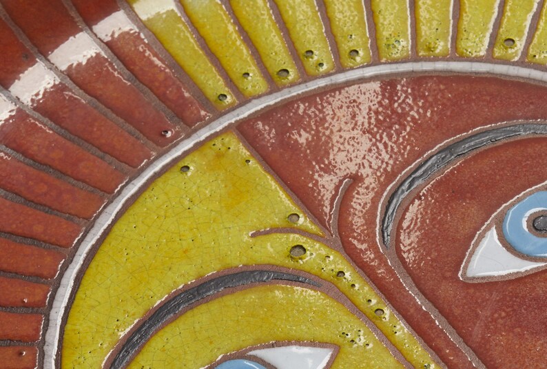 May include: Close-up of a ceramic sun face with yellow, red, and blue paint. The sun has a stylized face with two eyes and a nose. The sun is surrounded by yellow rays.