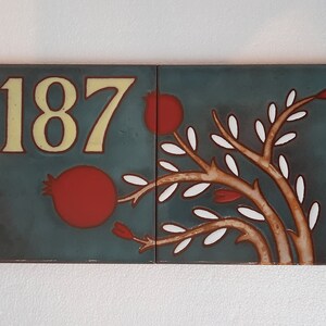 May include: Two blue ceramic tiles with a white background. The left tile has the number 1187 in yellow lettering. The right tile has a stylized tree with white leaves and red fruit.