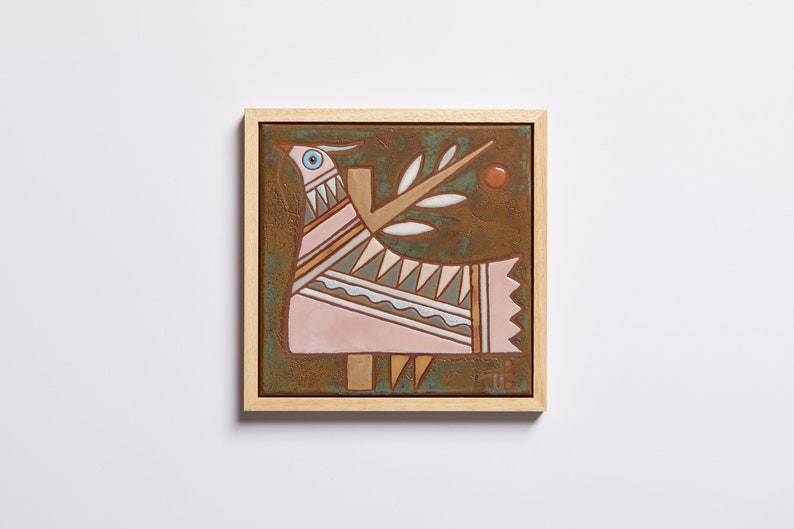May include: A framed ceramic tile with a stylized pink bird with geometric patterns and a white branch with leaves on a brown background.