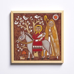 May include: A framed ceramic tile depicting a religious scene. The artwork features a woman holding a baby on a donkey, accompanied by a man with a staff. The color palette includes brown, red, yellow, and white, with a tree and birds in the background.