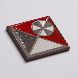 May include: A square ceramic tile with a geometric design. The tile is divided into four sections, each with a different color: red, white, gray, and black. The center of the tile features a black and white wheel with five spokes.