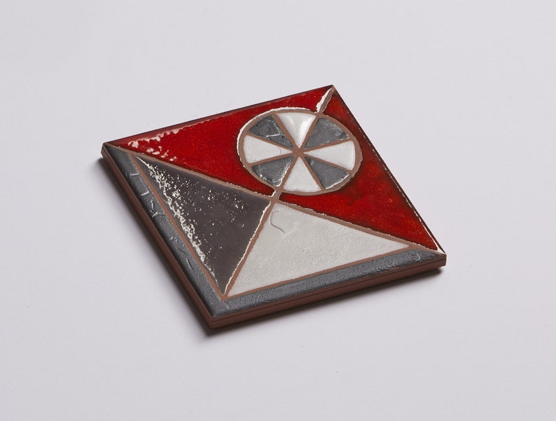 May include: A square ceramic tile with a geometric design. The tile is divided into four sections, each with a different color: red, white, gray, and black. The center of the tile features a black and white wheel with five spokes.