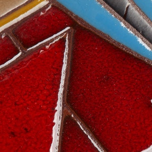 May include: Close-up of a red, blue, and white ceramic tile with a geometric pattern. The tile has a textured surface and is made of glazed ceramic.