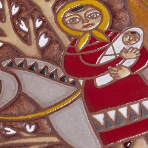 May include: A ceramic tile featuring a stylized illustration of a woman in a red dress and yellow head covering holding a baby wrapped in white. The woman is riding a gray horse with a yellow bridle. The background is brown with white leaf shapes.