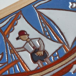 Peut inclure: Un carreau de c&eacute;ramique avec un motif peint &agrave; la main d'un voilier avec une seule voile, une personne &agrave; bord et deux oiseaux volant au-dessus. Le carreau est encadr&eacute; de bois et pr&eacute;sente une palette de couleurs bleue, blanche, orange et marron.