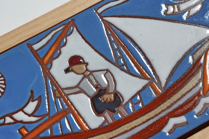 Peut inclure: Un carreau de c&eacute;ramique avec un motif peint &agrave; la main d'un voilier avec une seule voile, une personne &agrave; bord et deux oiseaux volant au-dessus. Le carreau est encadr&eacute; de bois et pr&eacute;sente une palette de couleurs bleue, blanche, orange et marron.