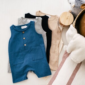 May include: Four sleeveless baby onesies in different colors and patterns. The onesies are made of a soft, lightweight fabric. The onesies are laid out on a white surface. The onesies are a light blue, a light brown, a black, and a gray with white stripes.