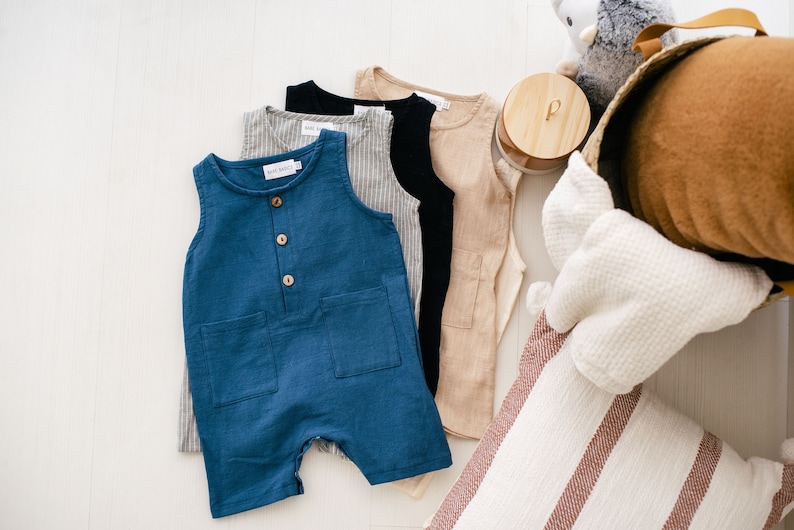 May include: Four sleeveless baby onesies in different colors and patterns. The onesies are made of a soft, lightweight fabric. The onesies are laid out on a white surface. The onesies are a light blue, a light brown, a black, and a gray with white stripes.
