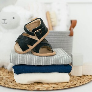 May include: A stack of baby clothes in grey, blue and white with a pair of black leather baby sandals on top.