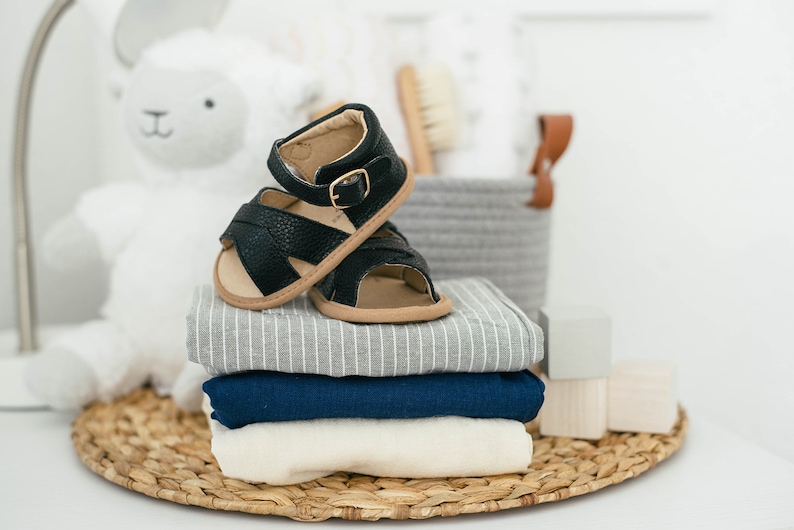 May include: A stack of baby clothes in grey, blue and white with a pair of black leather baby sandals on top.