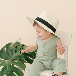 May include: A baby wearing a green jumpsuit and a white hat with a black band sits on a white chair. A large green leaf plant is in the background.