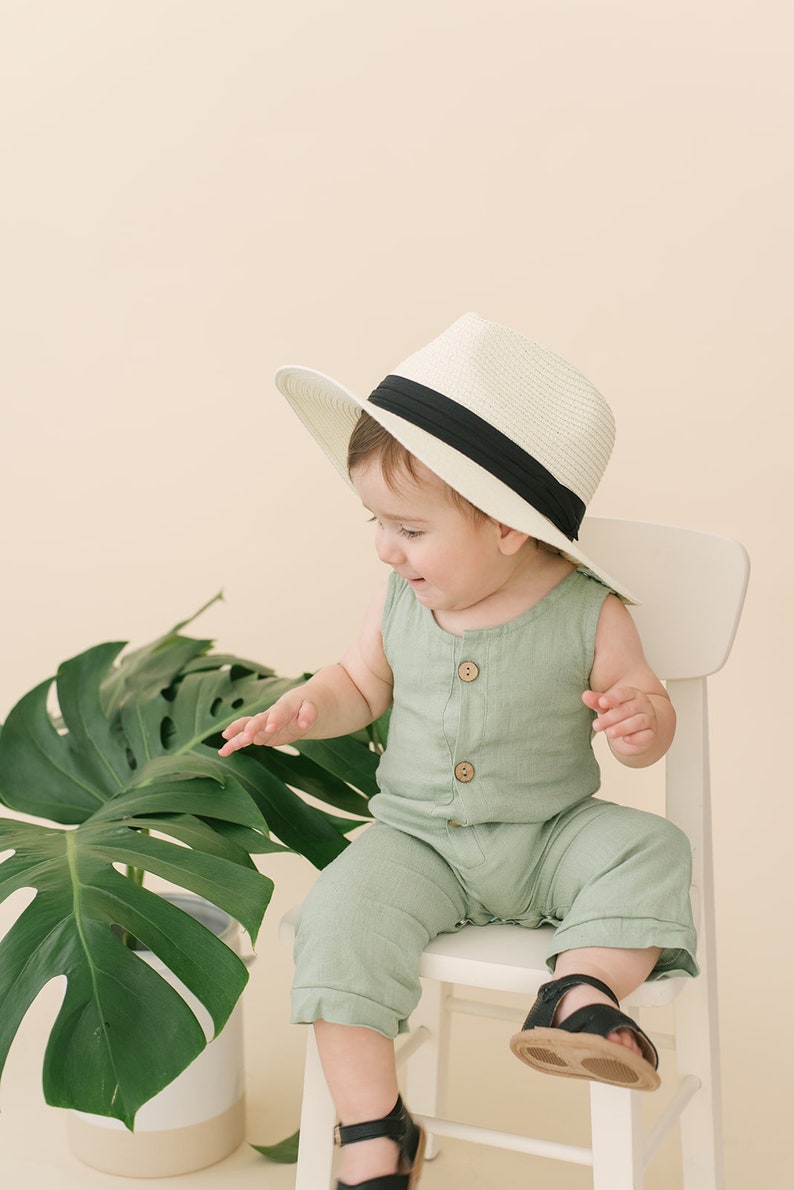 May include: A baby wearing a green jumpsuit and a white hat with a black band sits on a white chair. A large green leaf plant is in the background.