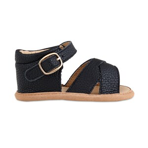 May include: Black leather sandals with a gold buckle and a criss-cross strap design. The sandals have a brown sole.