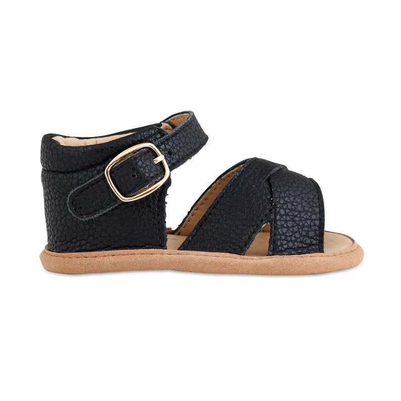 May include: Black leather sandals with a gold buckle and a criss-cross strap design. The sandals have a brown sole.